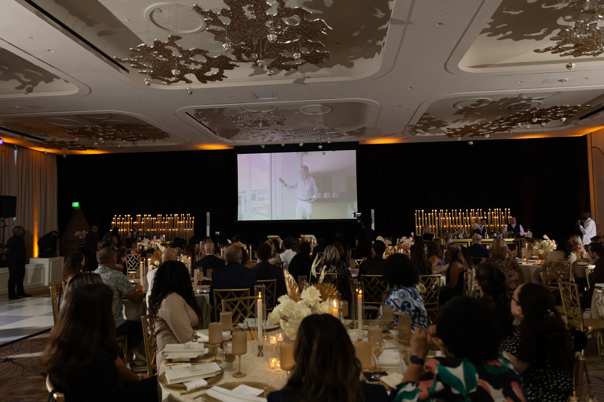 Colin Cowie NACE South Florida gala, full ballroom during keynote program, audience at gold chiavari chairs, Solene candle arrays flanking screen, EventPro event production Pier 66 Fort Lauderdale