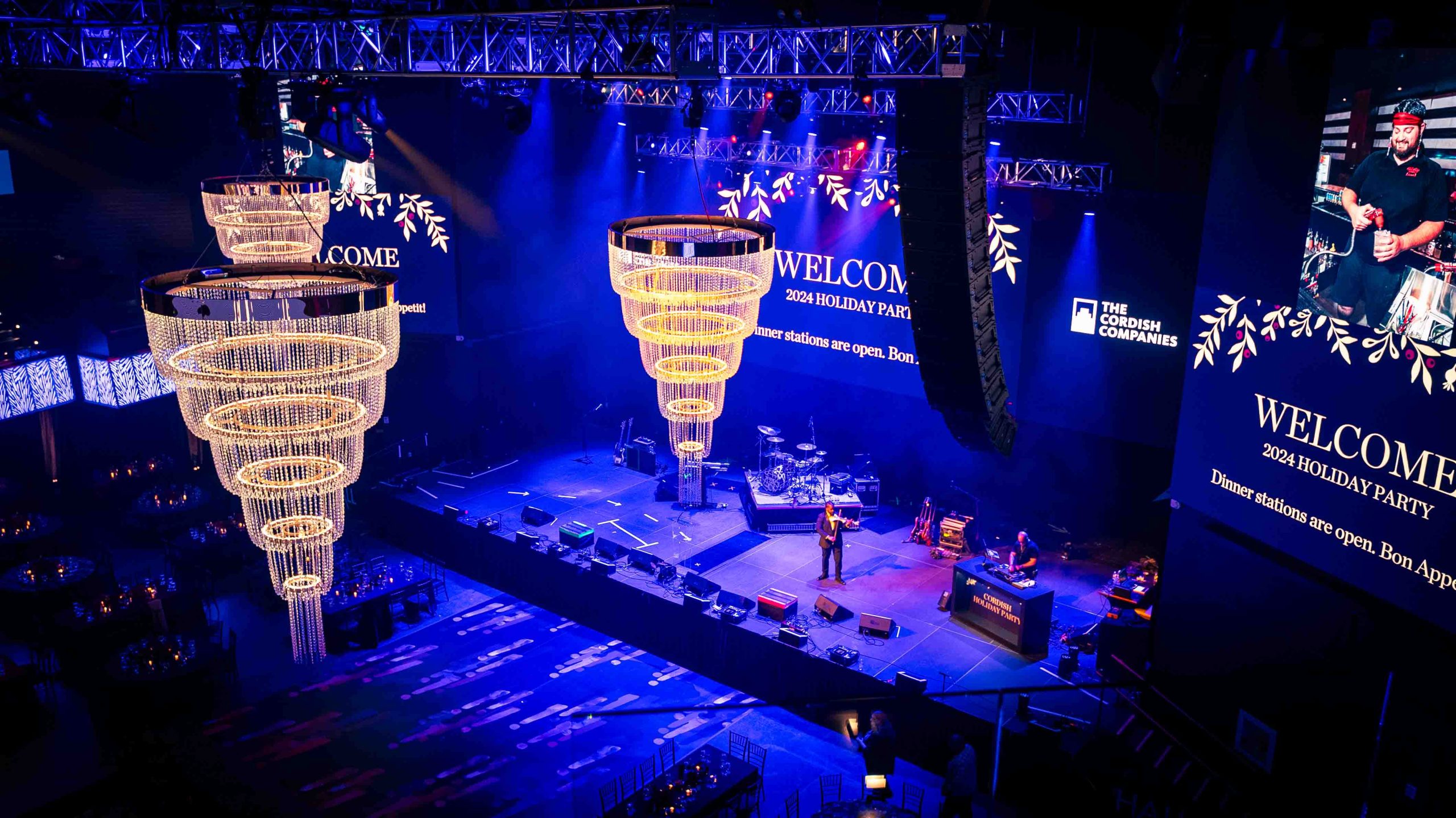 Performer addresses the room from stage, warm gold chandeliers overhead during dinner at the Cordish Companies 2024 Holiday Party, EventPro production
