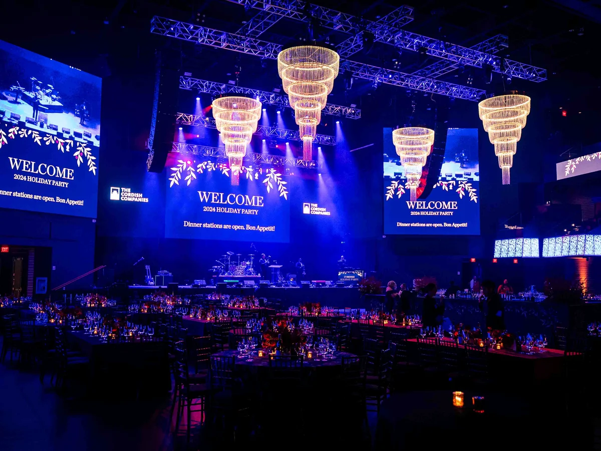 Gold ring chandeliers hung over corporate holiday party with blue wash lighting at large venue by Event Pro