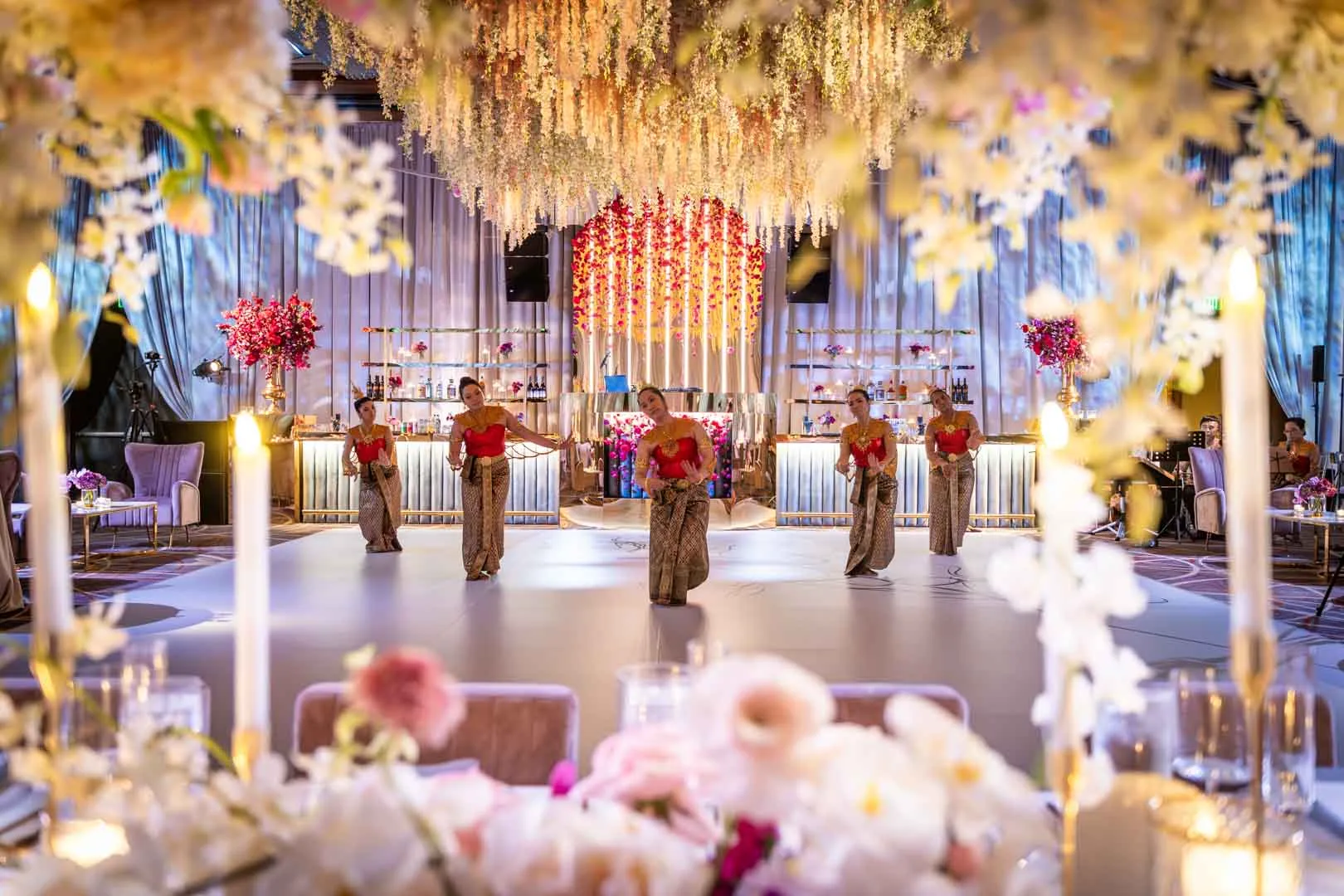 Wide reception view through floral centerpieces at Kam and Kay wedding Four Seasons Baltimore by Thai dancers on EventPro custom dance floor, floral ceiling and bar behind