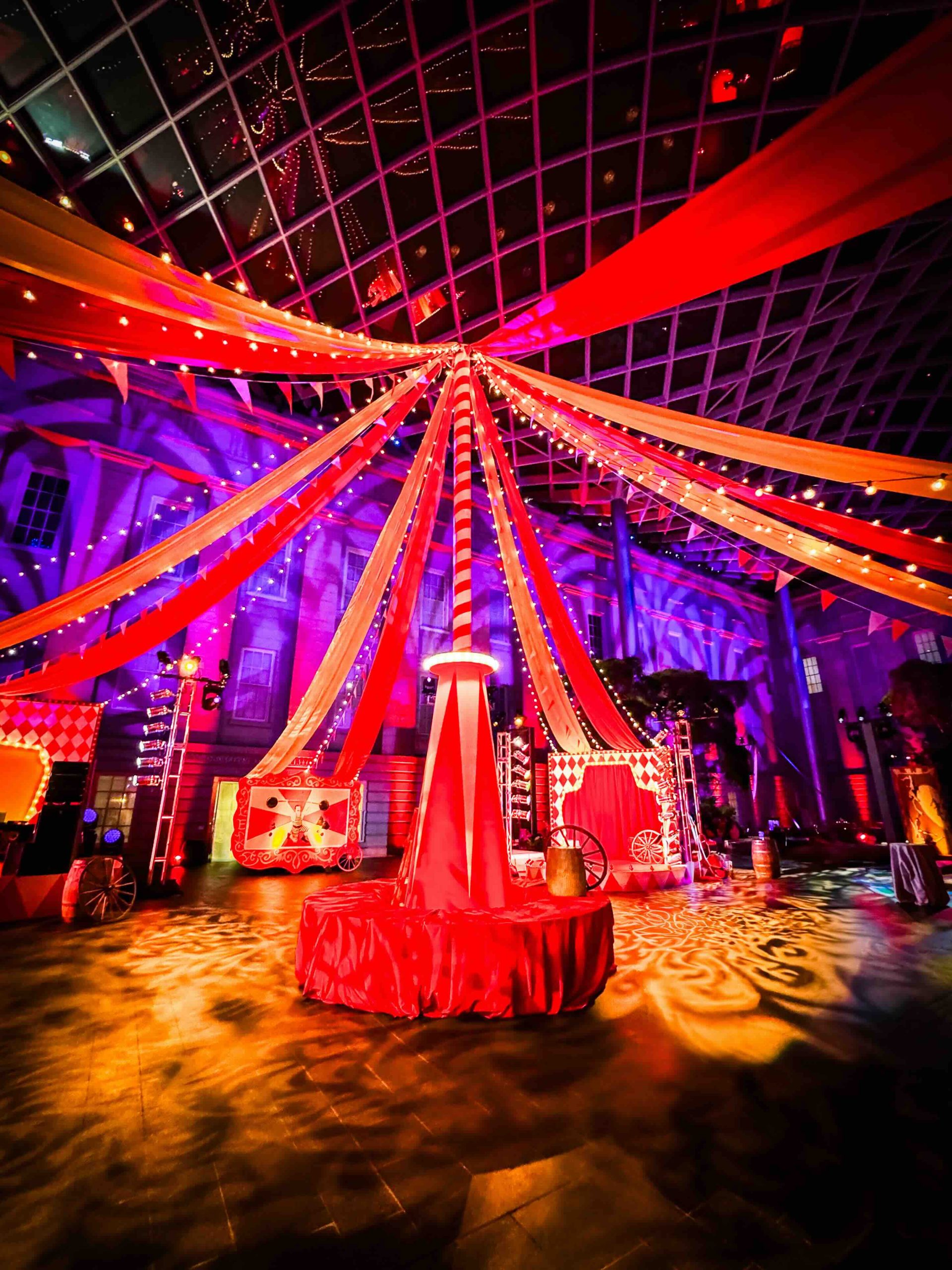 Looking up from the freestanding custom circus center pole, red and orange fabric canopy radiating to the National Portrait Gallery glass ceiling, EventPro DC Mardi Gras 2025