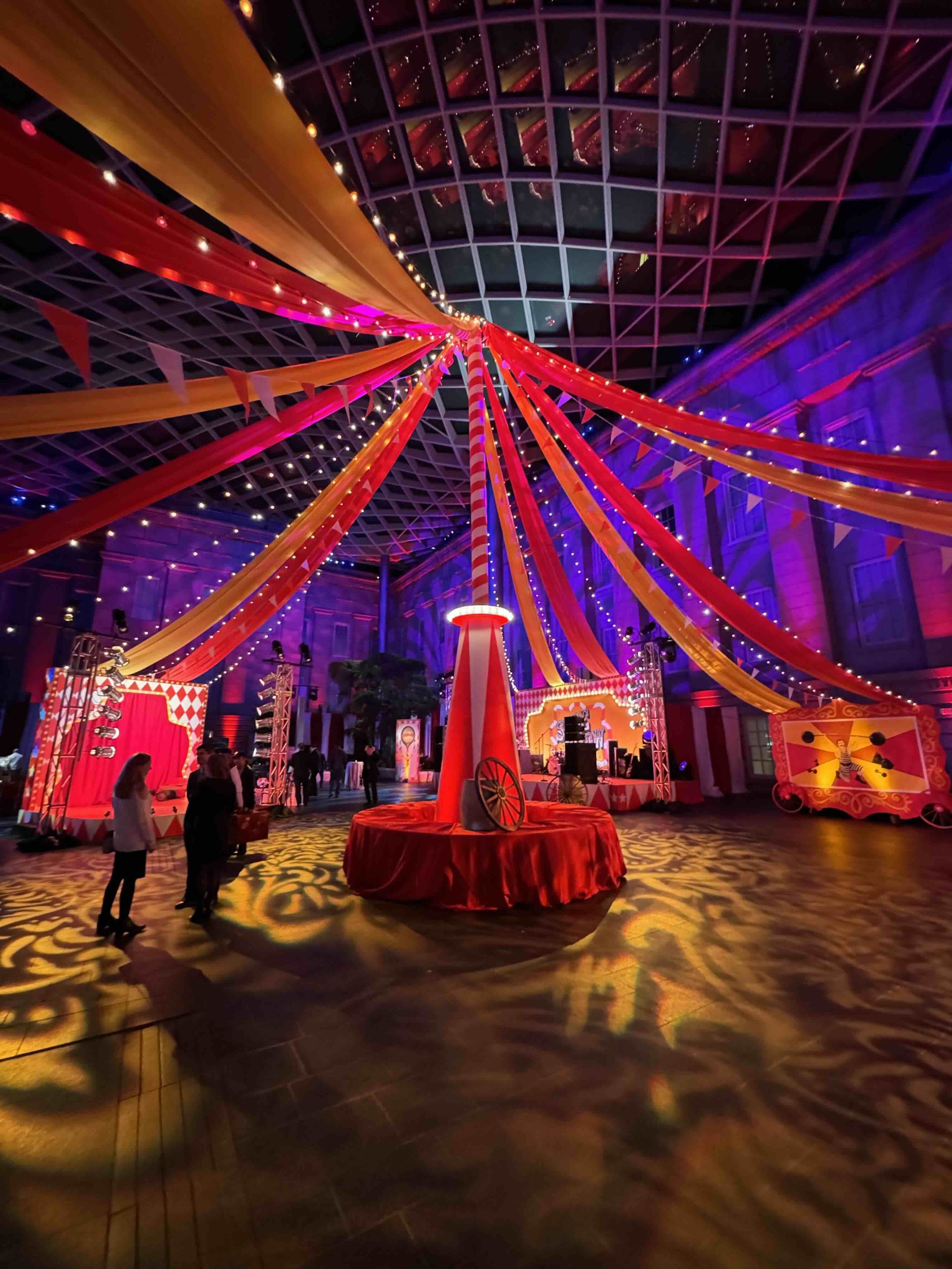 Full-room portrait from the entrance, freestanding circus center pole, fabric canopy, and carnival environment in the National Portrait Gallery Atrium, EventPro DC Mardi Gras 2025
