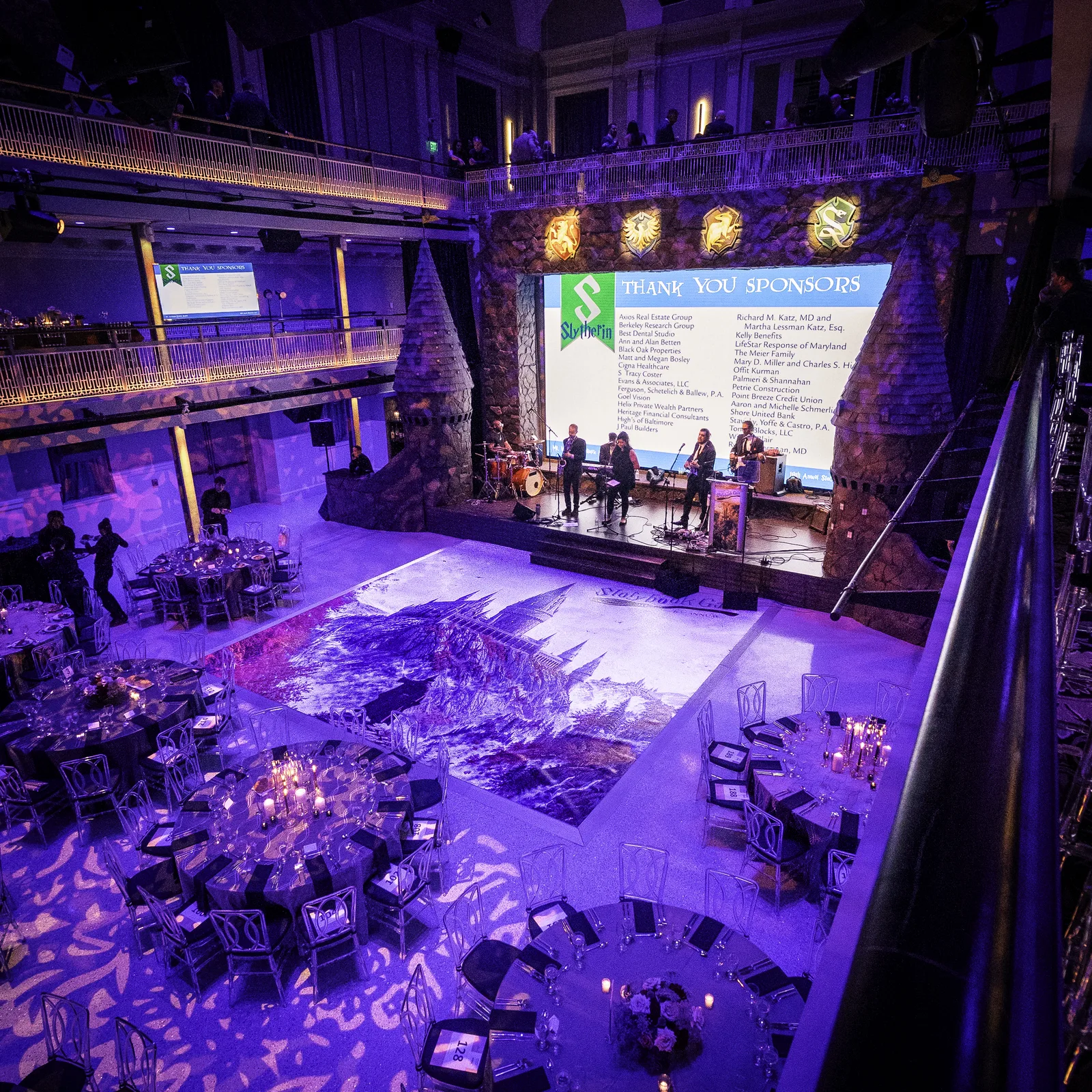 Overhead view during the program, printed Hogwarts castle floor wrap on dance floor, live band on stage behind castle arch set, Thank You Sponsors screen, EventPro production