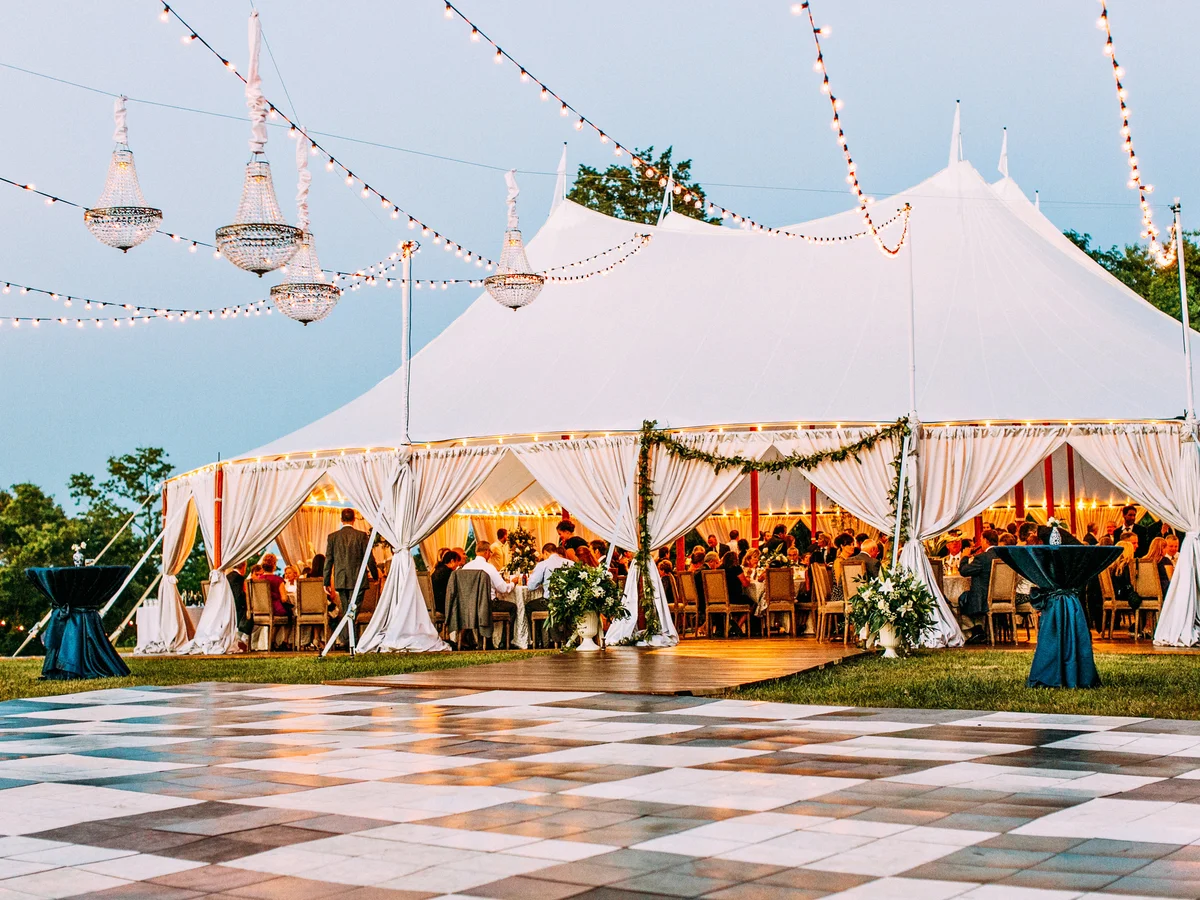 String lights and chandeliers draped over outdoor tented waterfront wedding at dusk by Event Pro