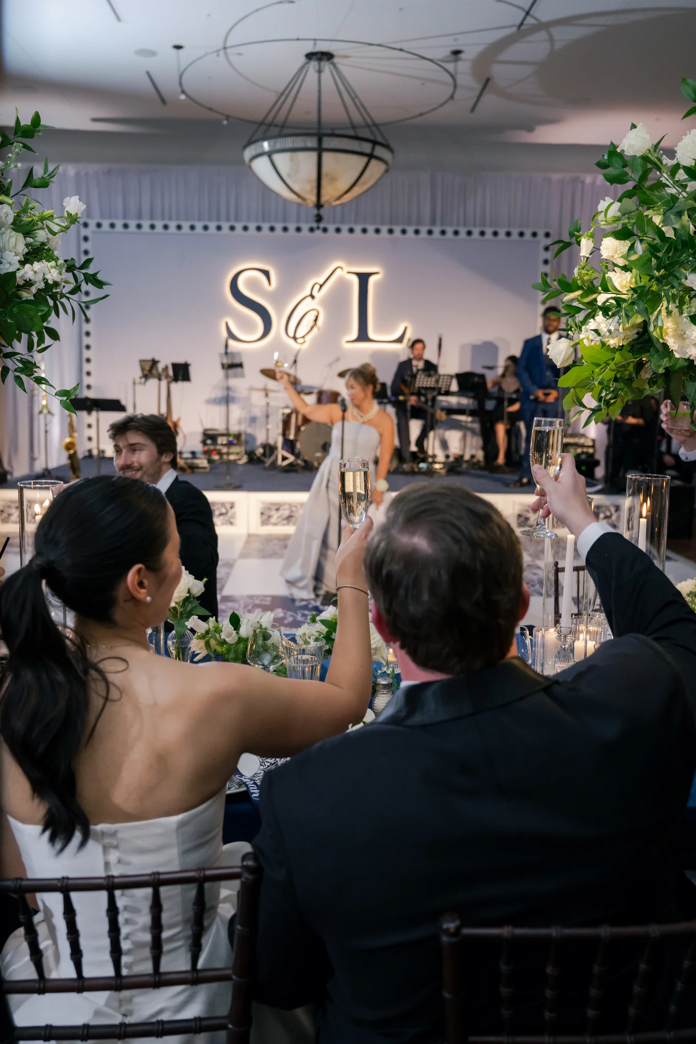 Champagne toast at Brittland Estates wedding, couple raising glasses at sweetheart table, EventPro LED-backlit S&L monogram and custom stage visible behind, Maryland wedding production