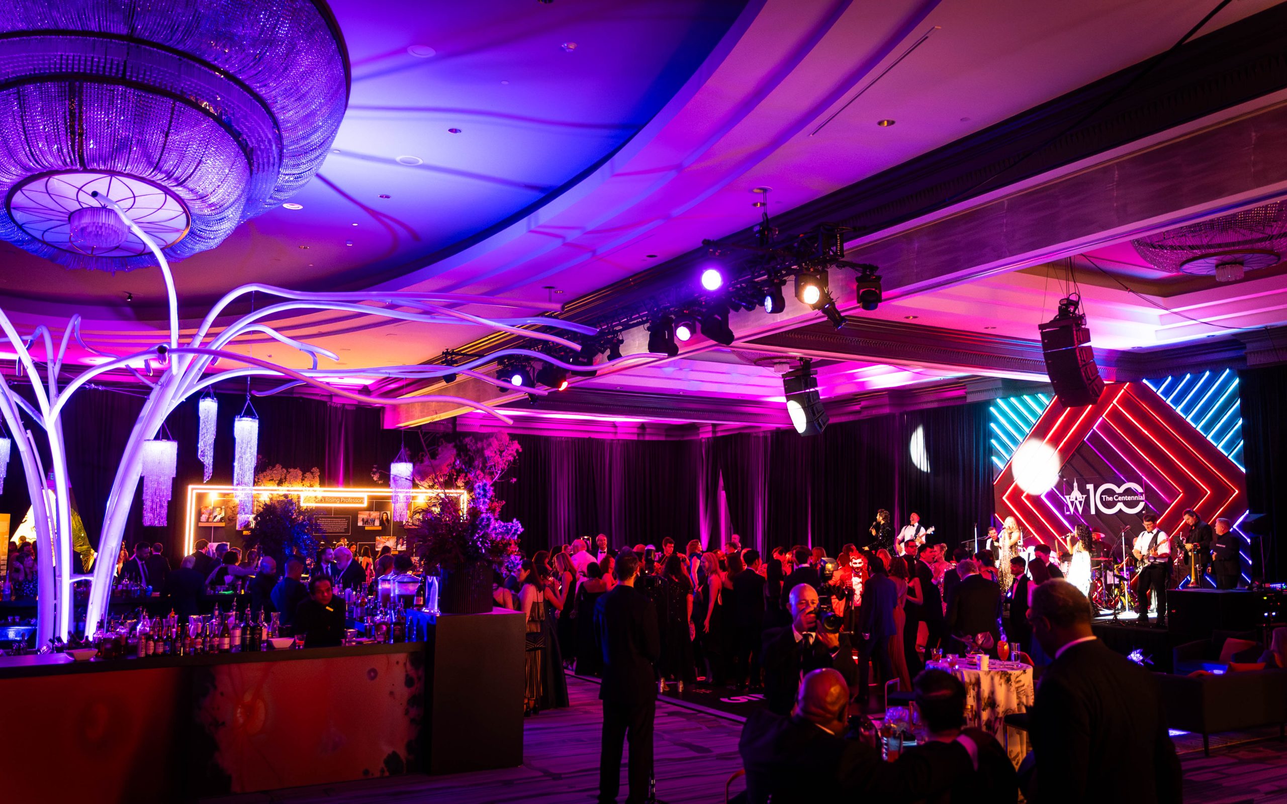 Full ballroom during the concert, branching white tree sculpture lit in blue, Downtown Band on stage with neon diamond backdrop, guests dancing, Wilmer Eye Institute Centennial Gala, EventPro